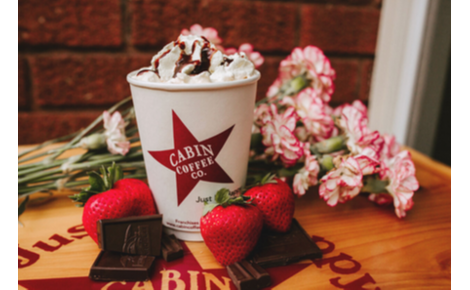 Cabin Coffee Cup with a delicious drink sitting on a table with flowers behind it. Strawberries and chocolate sit in front of the drink. 