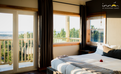 hotel room showing the bed with a throw blanket folded at the foot. Large French doors with windows looks out onto the deck and the ocean. 